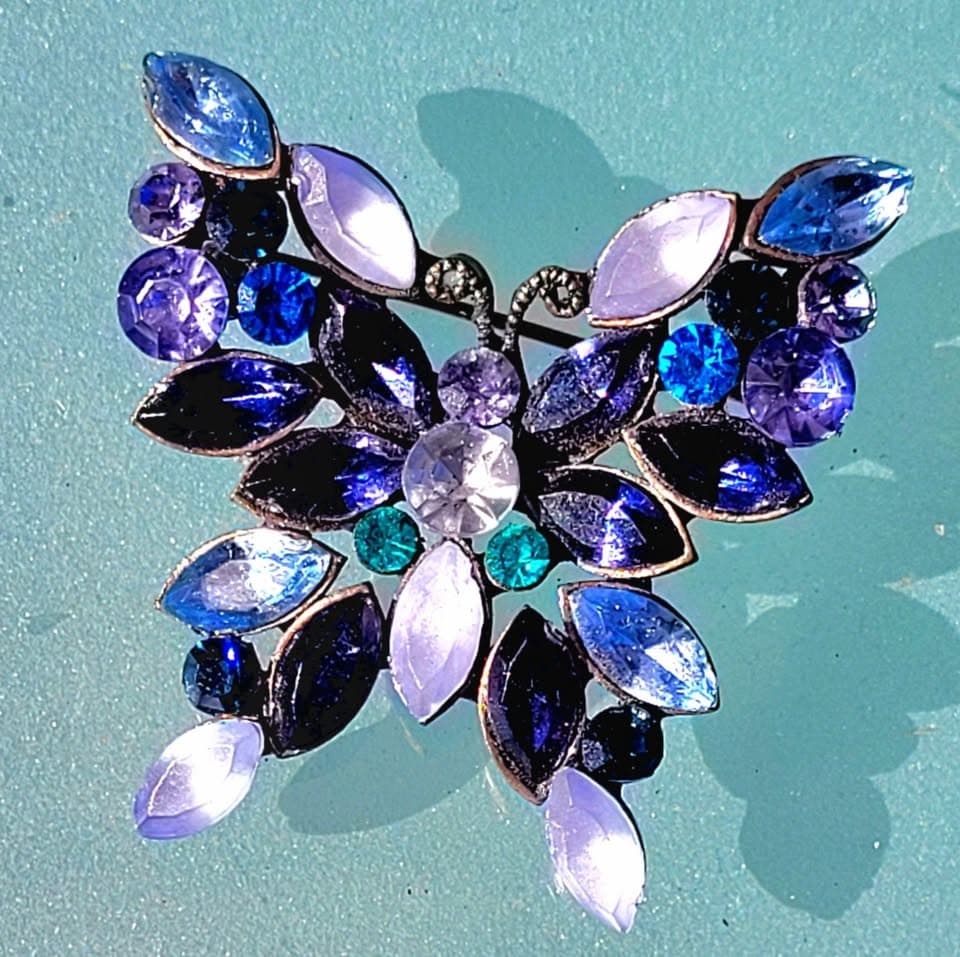 A butterfly-shaped brooch set with blue, amethyst, and icy-lavender rhinestones on a teal ground