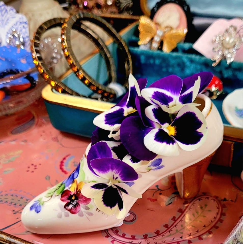 A crowded display of a hand-painted porcelain shoe trimmed with fresh pansies, rhinestone bangles, and jewelry on teal velvet trays over a coral paisley cloth