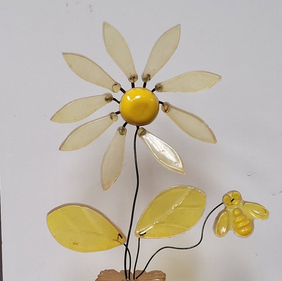 A folk-art flower sculpture with yellow acrylic petals, a daisy center, and a small bee on a wire stem